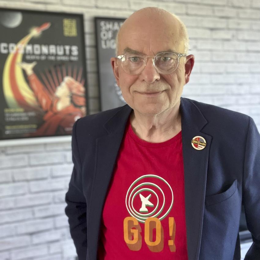 A picture of physicist Mark Wrigley pictured in a white brick hallway. He's an older gentleman wearing glasses and dressed in a red t-shirt and blue blazer. 