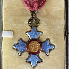A medal designating the award of Commander of the Order of the British Empire. The medal has a red ribbon, a metal crown and four blue arms around two figures with crowns on a central disc.