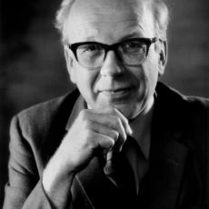 A black and white portrait of Professor Sir Francis Graham Smith. He rests his chin on his hand and smiles at the camera. He is wearing glasses, a shirt, tie and blazer.