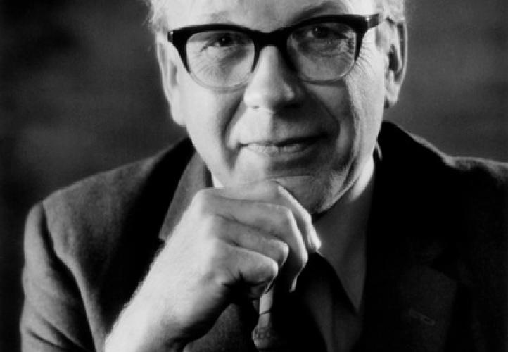 A black and white portrait of Professor Sir Francis Graham Smith. He rests his chin on his hand and smiles at the camera. He is wearing glasses, a shirt, tie and blazer.