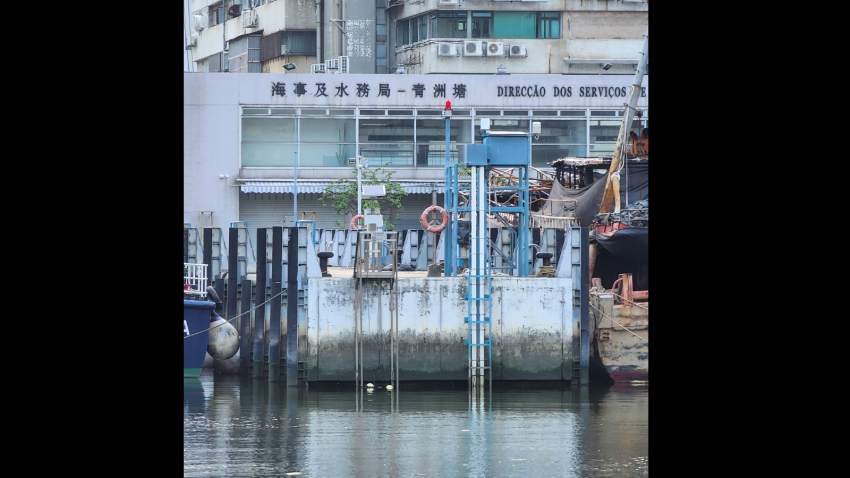 The SDV tide gauge at Macau. A wall with very obvious water markings. Behind the wall are several buildings. Water beneath the wall and markings is dark and still.