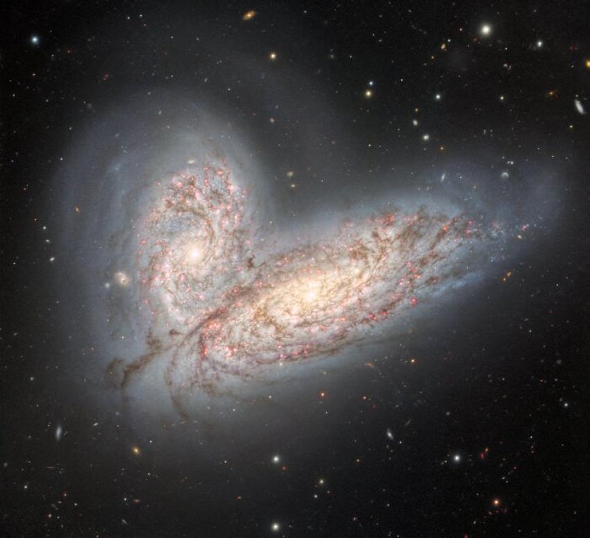 An image of two merging galaxies. Both are light blue and cream in colour. Faint stars are seen in the background.