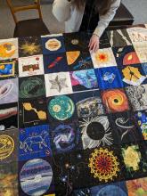 The bicentenary patchwork quilt laid out on a table, with a women working on stitching it together. It features embroidered squares depicting galaxies, planets and other celestial objects. 